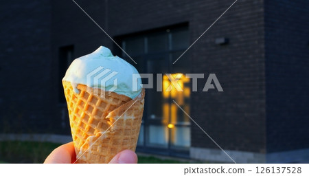 Light green pistachio ice cream in a crispy waffle cone is melting slightly while being held in front of a dark modern building with glass windows reflecting warm interior lights Light green pistachio ice cream in a crispy waffle cone is melting slightly while being held in front of a dark modern building with glass windows reflecting warm interior lights 126137528