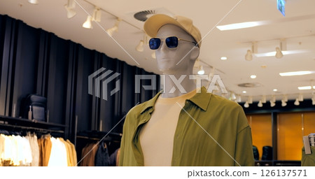 Fashionable mannequin presenting a casual summer outfit including a green shirt, beige t shirt, beige cap and sunglasses, displayed in a clothing store. Concept fashion clothes 126137571