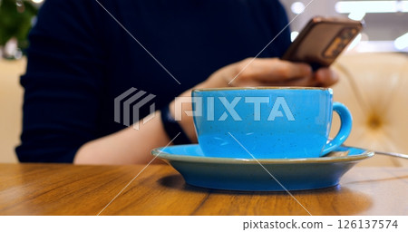 Multitasking professional relaxing during break, sipping coffee while scrolling smartphone wooden table in cozy cafe environment, demonstrating modern digital lifestyle. Concept of coffee consumption 126137574