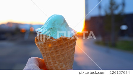 Creamy light green pistachio ice cream nestled in waffle cone, gleaming against blurred urban sunset street with golden evening glow and soft bokeh background 126137624