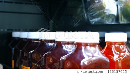 Red juice in clear plastic bottles with white caps sits on a refrigerated shelf, likely in a grocery store or supermarket, with additional packaged goods visible in the background 126137625
