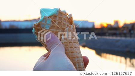 Delicate hand holding melting pistachio ice cream in waffle cone, highlighting sunset river scenery with golden evening light and soft color reflections Delicate hand holding melting pistachio ice cream in waffle cone, highlighting sunset river scenery with golden evening light and soft color reflections 126137637