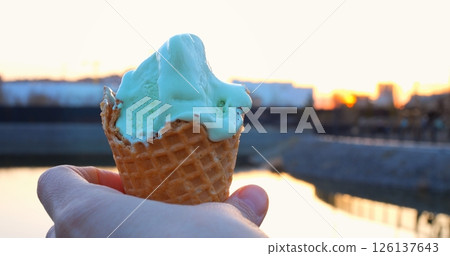 Delicious pistachio ice cream melts slowly down a crispy waffle cone, held by a hand in the warm glow of a beautiful sunset, with blurred urban background Delicious pistachio ice cream melts slowly down a crispy waffle cone, held by a hand in the warm glow of a beautiful sunset, with blurred urban background 126137643