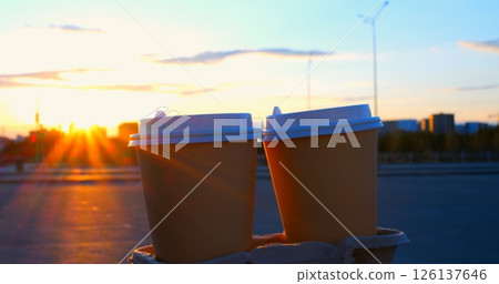 Sunlight shines between two disposable coffee cups on a cardboard tray, creating a starburst effect, with a blurred urban background at sunset Sunlight shines between two disposable coffee cups on a cardboard tray, creating a starburst effect, with a blurred urban background at sunset 126137646