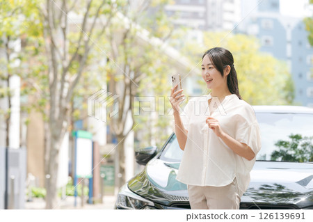 A young woman operating a smartphone in front of a car 126139691