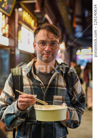 Russian male tourist eating crab and pork dumplings with chopsticks on Yuyuan Old Street in Shanghai, wearing glasses and a plaid shirt Russian male tourist eating crab and pork dumplings with chopsticks on Yuyuan Old Street in Shanghai, wearing glasses and a plaid shirt 126140634
