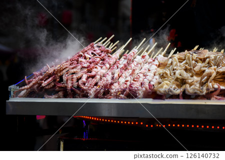 Night street food market in Shanghai, China. Grilled seafood skewers octopus and tentacles sizzling on fire. Dark, atmospheric vibe. Night street food market in Shanghai, China. Grilled seafood skewers octopus and tentacles sizzling on fire. Dark, atmospheric vibe. 126140732