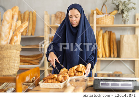 Girl in traditional Muslim clothing selects and places fresh hot croissants in plastic bag Girl in traditional Muslim clothing selects and places fresh hot croissants in plastic bag 126143871