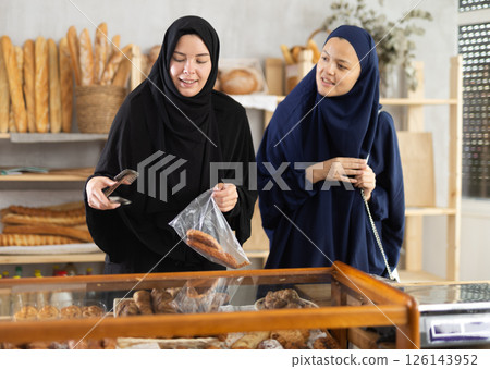 Female friends in Muslim attires choosing baked goods in bakery Female friends in Muslim attires choosing baked goods in bakery 126143952