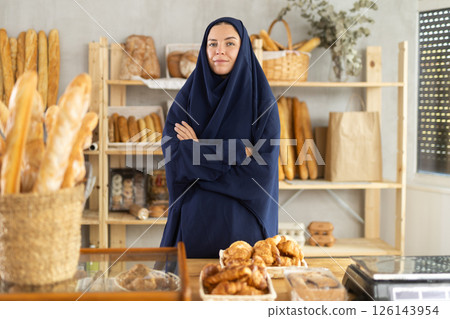 Muslim girl seller employee puts croissants in window, arranges display of goods at bakery. Muslim girl seller employee puts croissants in window, arranges display of goods at bakery. 126143954