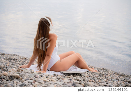 A woman is sitting on a beach with her headphones on. She is wearing a white bikini and a white shirt. The beach is rocky and the water is calm. The woman is enjoying her time by the water. A woman is sitting on a beach with her headphones on. She is wearing a white bikini and a white shirt. The beach is rocky and the water is calm. The woman is enjoying her time by the water. 126144191