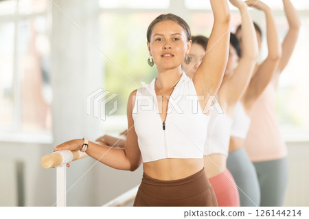 Group ballet rehearsal - young ballerinas stand in third position near ballet barre during group training in dance studio Group ballet rehearsal - young ballerinas stand in third position near ballet barre during group training in dance studio 126144214