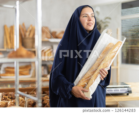 Young female customer in blue abaya holding baguettes in bakery Young female customer in blue abaya holding baguettes in bakery 126144283