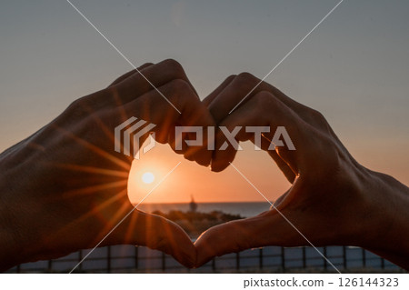 Sunset Heart Hands Beach - Silhouette of hands framing a sunset over the beach, forming a heart shape. 126144323