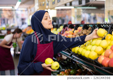 Female seller in hijab and an apron puts fresh apples on shelves in the grocery section of supermarket 126144398