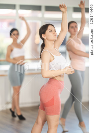 Warm-up sessions. Cheerful fit active young girl training various ballet poses during group classes with ballet dancers, rehearsal before performance in choreography studio Warm-up sessions. Cheerful fit active young girl training various ballet poses during group classes with ballet dancers, rehearsal before performance in choreography studio 126144446
