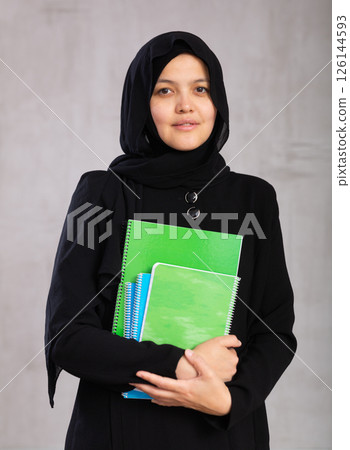 Portrait of young woman in hijab holding stack of notebooks Portrait of young woman in hijab holding stack of notebooks 126144593