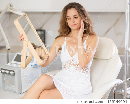 Young upset woman examining her face after cosmetic procedure Young upset woman examining her face after cosmetic procedure 126144694