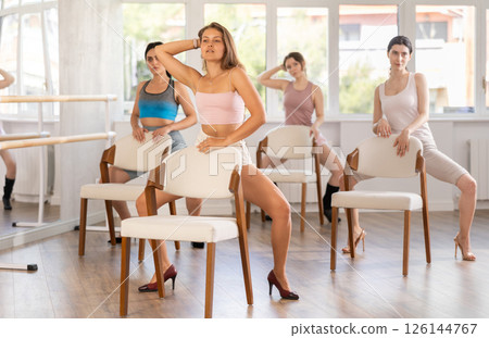 Leaning on chair seat with hand foot, young woman performs lap dance during group workout. 126144767