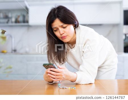 Upset young brunette standing in home kitchen with smartphone Upset young brunette standing in home kitchen with smartphone 126144787