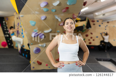 Positive slender europian girl in sportswear is standing near climbing wall in gym Positive slender europian girl in sportswear is standing near climbing wall in gym 126144817