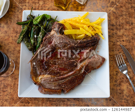 Grilled beef chop on bone, served with a vegetable side dish of fried potatoes and green padron peppers Grilled beef chop on bone, served with a vegetable side dish of fried potatoes and green padron peppers 126144987