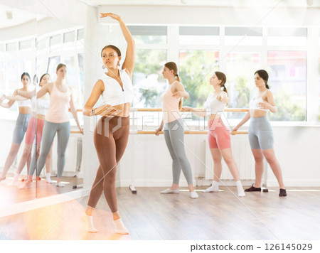 Young beautiful female ballet student demonstrating skills in ballet in dance studio. Group of young ballet dancers in background near ballet barre Young beautiful female ballet student demonstrating skills in ballet in dance studio. Group of young ballet dancers in background near ballet barre 126145029