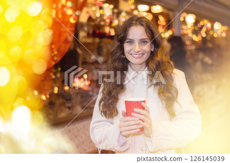 Cheerful woman with paper cup of hot tea walking at Christmas street fair Cheerful woman with paper cup of hot tea walking at Christmas street fair 126145039