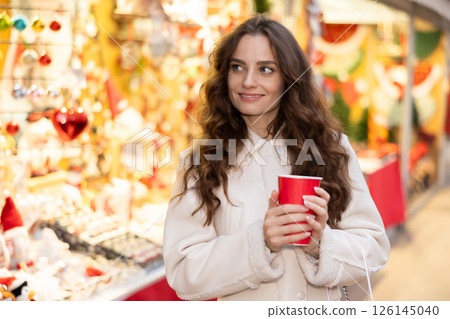 Cheerful woman with paper cup of hot tea walking at Christmas street fair Cheerful woman with paper cup of hot tea walking at Christmas street fair 126145040