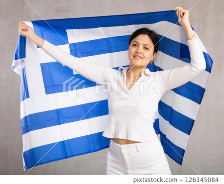 Middle-aged positive woman in casual clothes hold unfurled flag of Greece in hands raised above head 126145084
