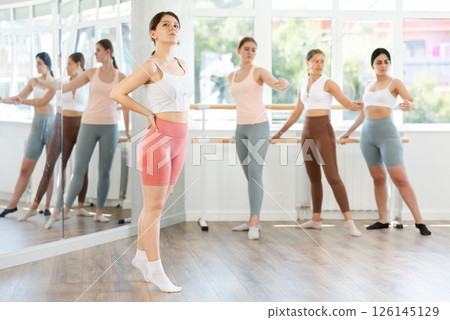 Young beautiful female ballet student demonstrating skills in ballet in dance studio. Group of young ballet dancers in background near ballet barre 126145129