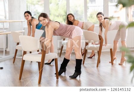 Leaning on chair seat with hand foot, young woman performs lap dance during group workout. 126145132
