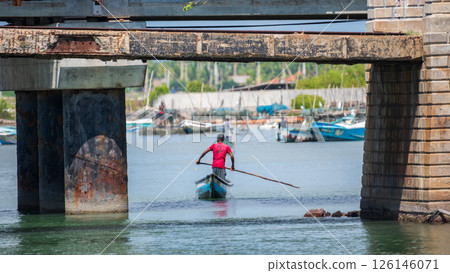A man in a red shirt rows a small traditional boat under an old, weathered bridge in Mannar, Sri Lanka. Fishing harbor in the background A man in a red shirt rows a small traditional boat under an old, weathered bridge in Mannar, Sri Lanka. Fishing harbor in the background 126146071