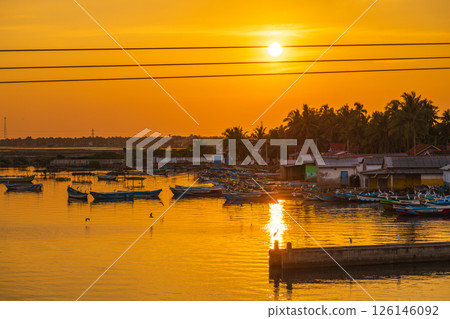 A glowing orange sunset and the quiet boatyard in warm light, with calm waters reflecting the golden sky. Fishing boats rest peacefully on the water near the shore A glowing orange sunset and the quiet boatyard in warm light, with calm waters reflecting the golden sky. Fishing boats rest peacefully on the water near the shore 126146092