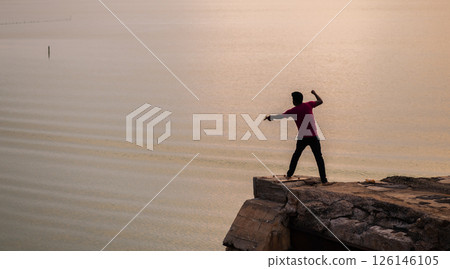 A lone fisherman stands at the edge of a rocky platform, casting his line into the calm waters of the Mannar lagoon in Sri Lanka at sunset, silhouetted against the soft golden hues A lone fisherman stands at the edge of a rocky platform, casting his line into the calm waters of the Mannar lagoon in Sri Lanka at sunset, silhouetted against the soft golden hues 126146105