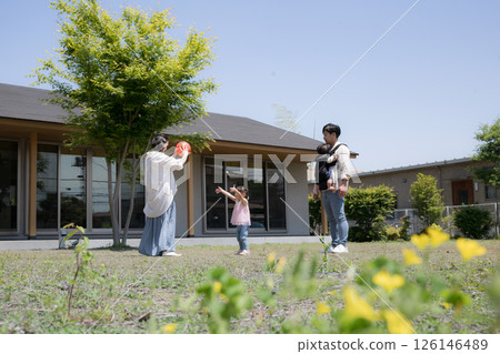 Happy family image of a family and children playing with a ball in front of the garden of a single-story house, blurred greenery in front 126146489