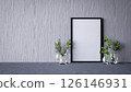 A minimalist setting featuring a blank frame against a textured wall, complemented by glass vases containing greenery. 126146931