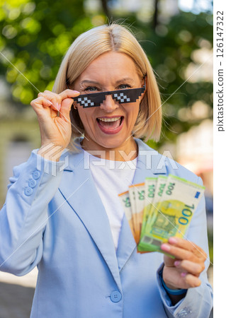 Happy rich mature business woman putting on thug life sunglasses showing euro money on city street Happy rich mature business woman putting on thug life sunglasses showing euro money on city street 126147322