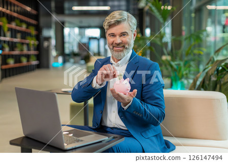 Happy mature businessman save money using pink piggybank putting dollar cast inside in office lobby 126147494
