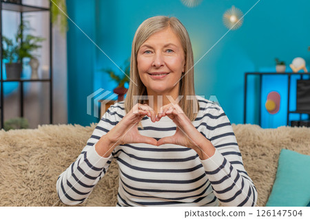 Mature woman making heart gesture on sofa, expressing love and gratitude, showing affection at home Mature woman making heart gesture on sofa, expressing love and gratitude, showing affection at home 126147504