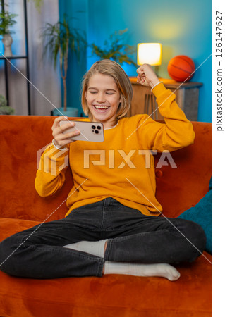 Teenager boy celebrating victory after winning mobile game on smartphone joyful jump on sofa at home 126147627