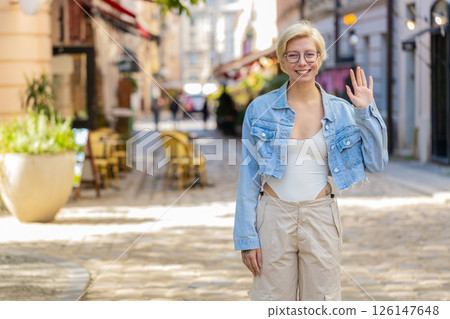 Cheerful young blonde woman smiling friendly at camera waving hands gesturing on city urban street Cheerful young blonde woman smiling friendly at camera waving hands gesturing on city urban street 126147648