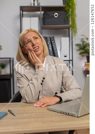 Upset mature businesswoman in suit suffering from toothache while using laptop at home office desk 126147652