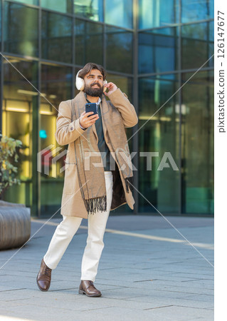 Happy young man putting on headphones listening favorite energetic music on smartphone on street Happy young man putting on headphones listening favorite energetic music on smartphone on street 126147677