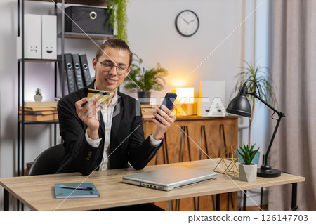 Happy young businessman using bank credit card and smartphone for online shopping at home office Happy young businessman using bank credit card and smartphone for online shopping at home office 126147703