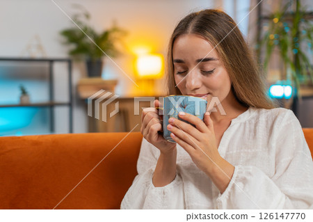 Smiling young woman sitting on sofa drinking cup of warm coffee or herbal tea in morning at home 126147770