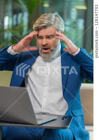 Sad businessman using laptop loses becoming shocked sudden lottery results in modern hotel lobby Sad businessman using laptop loses becoming shocked sudden lottery results in modern hotel lobby 126147783