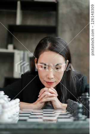 Woman deeply focused on a chess game, contemplating her next move in a modern room with a bookshelf. Woman deeply focused on a chess game, contemplating her next move in a modern room with a bookshelf. 126148309