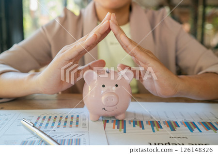 A woman shields a piggy bank with her hands, symbolizing financial security, amidst business reports and charts. A woman shields a piggy bank with her hands, symbolizing financial security, amidst business reports and charts. 126148326