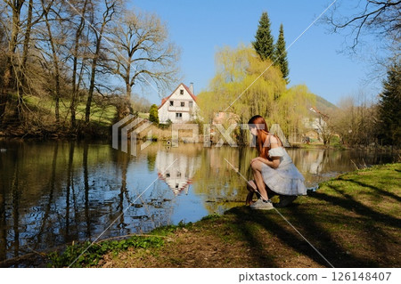 Young woman in white dress sitting by tranquil lake reflecting traditional European house and spring trees under clear blue sky in rural countryside landscape. High quality photo 126148407